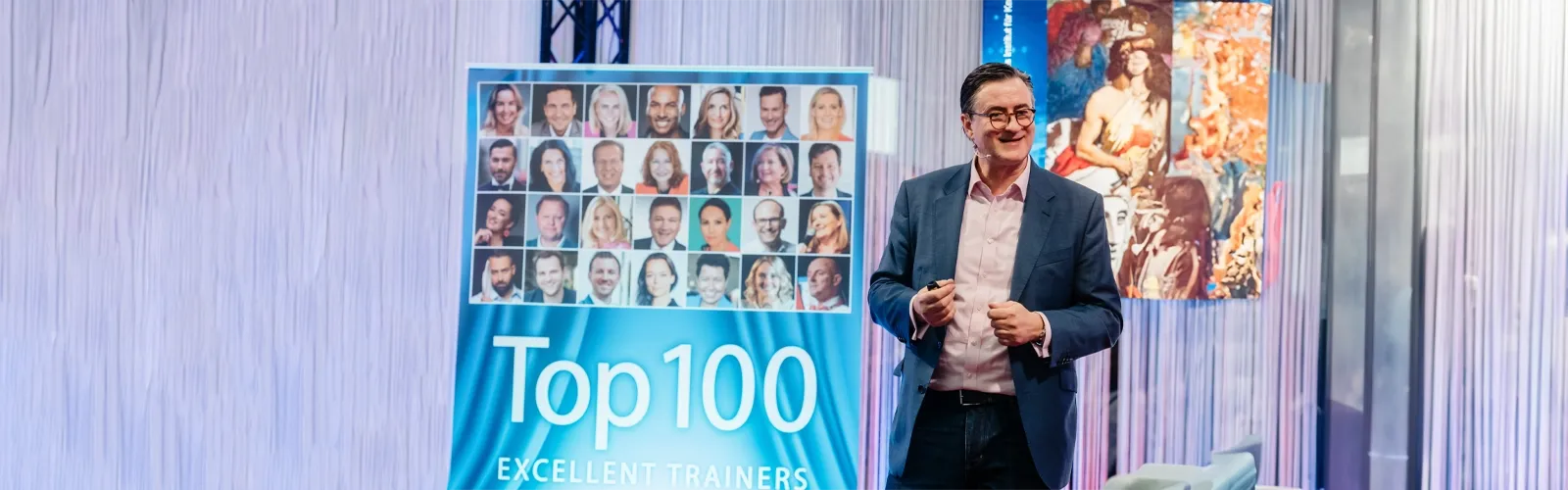 Medientrainer Dr. Nikolai A. Behr im Anzug steht auf der Bühne neben einem bunten Display, das Kopfbilder verschiedener Personen und den Text "Top 100 Excellent Trainers" zeigt. Im Hintergrund sind Vorhänge und ein Bildausschnitt eines Gemäldes zu sehen.