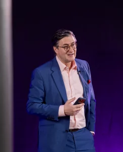 Redner Dr. Nikolai A. Behr in einem blauen Anzug und einem rosa Hemd steht vor einem dunklen Hintergrund, hält ein Telefon und lächelt. Er trägt eine Brille und hat ein Mikrofon an seinem Revers befestigt.