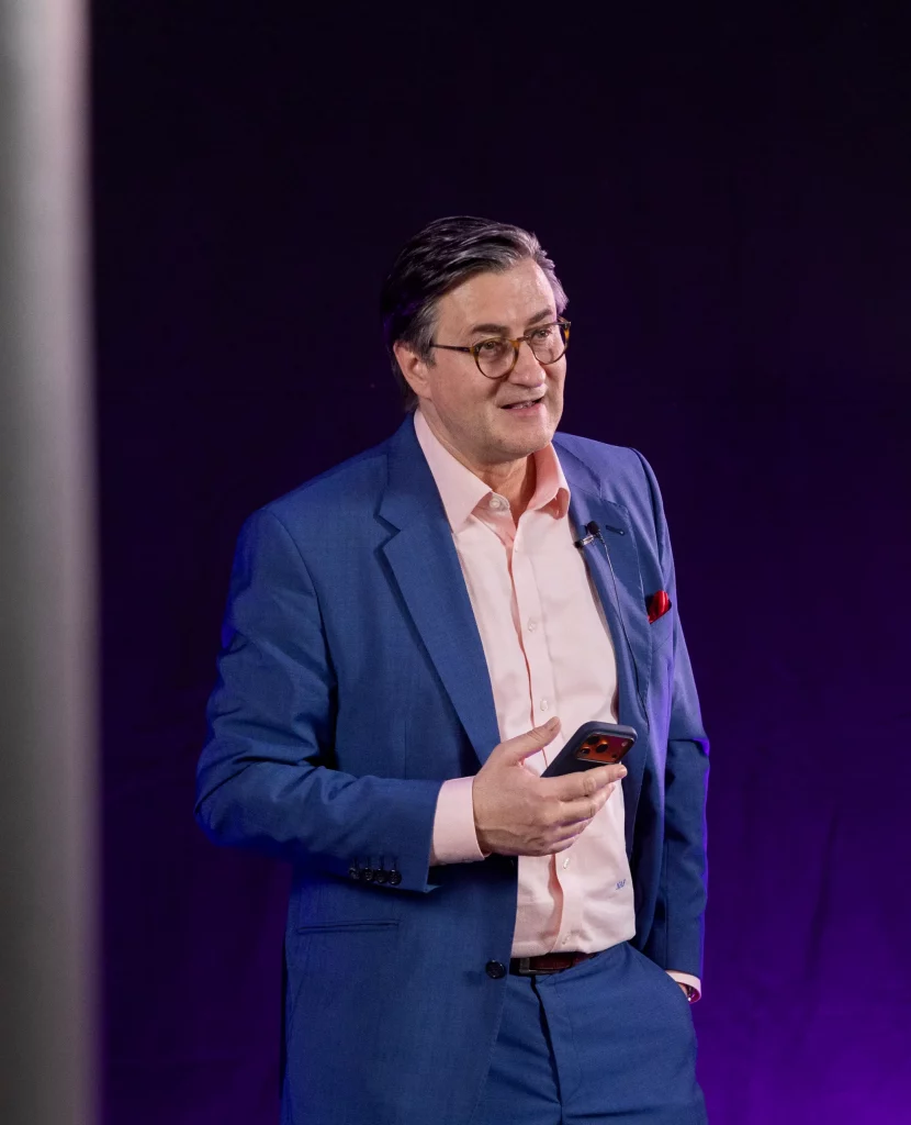 Redner Dr. Nikolai A. Behr in einem blauen Anzug und einem rosa Hemd steht vor einem dunklen Hintergrund, hält ein Telefon und lächelt. Er trägt eine Brille und hat ein Mikrofon an seinem Revers befestigt.