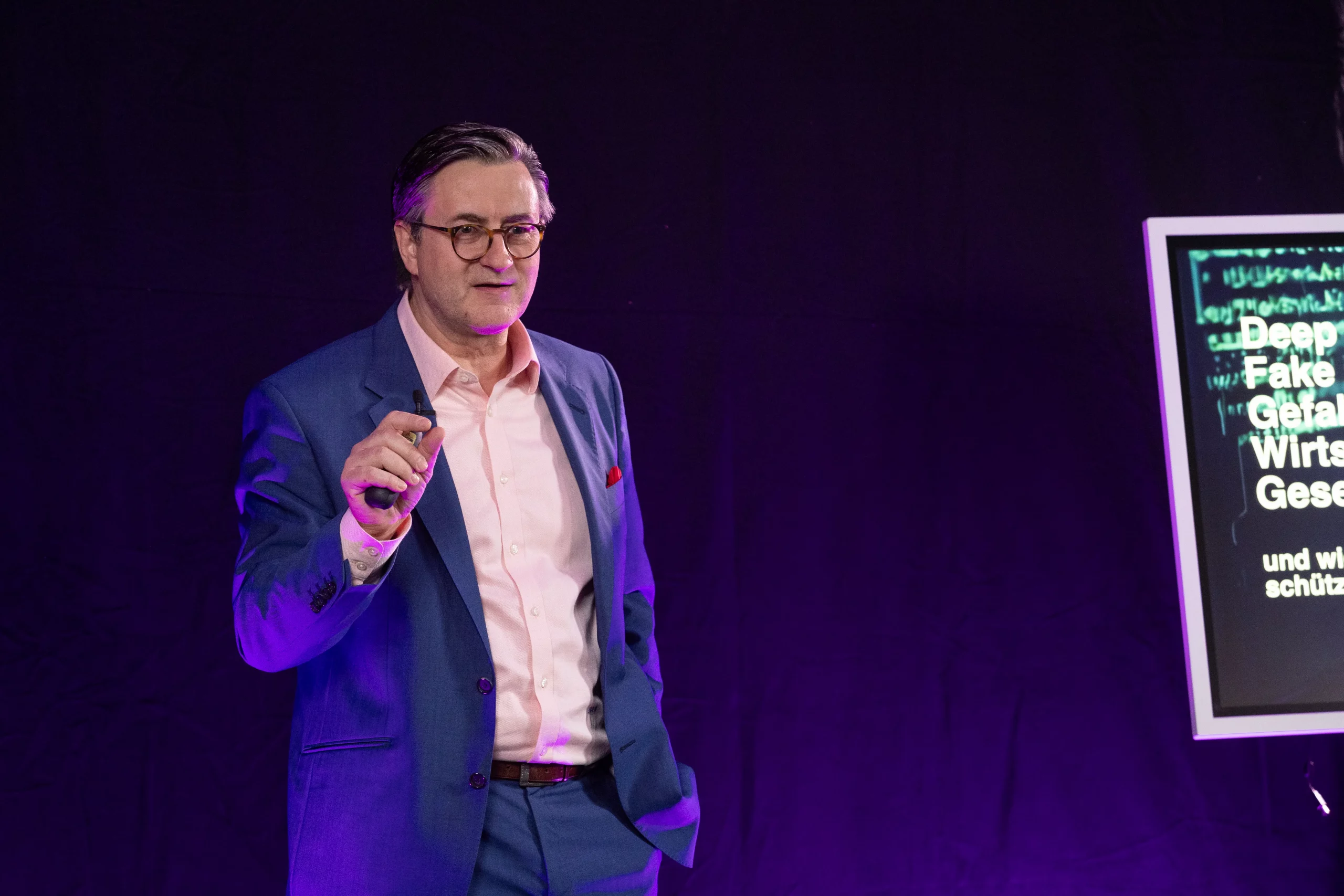 Redner Dr. Nikolai A. Behr in blauem Anzug und rosa Hemd steht vor einem dunklen Hintergrund, spricht und gestikuliert, während neben ihm ein Bildschirm mit teilweise sichtbarem Text angezeigt wird.