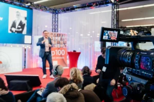 Eine Kamera filmt den Redner Dr. Nikolai A. Behr auf der Bühne einer Konferenz. Die Zuhörer sitzen auf Stühlen, einige mit Kopfhörern, während im verschwommenen Hintergrund ein Bildschirm mit dem Foto einer Person und einem Banner zu sehen ist. Die Szene ist lebendig und professionell.