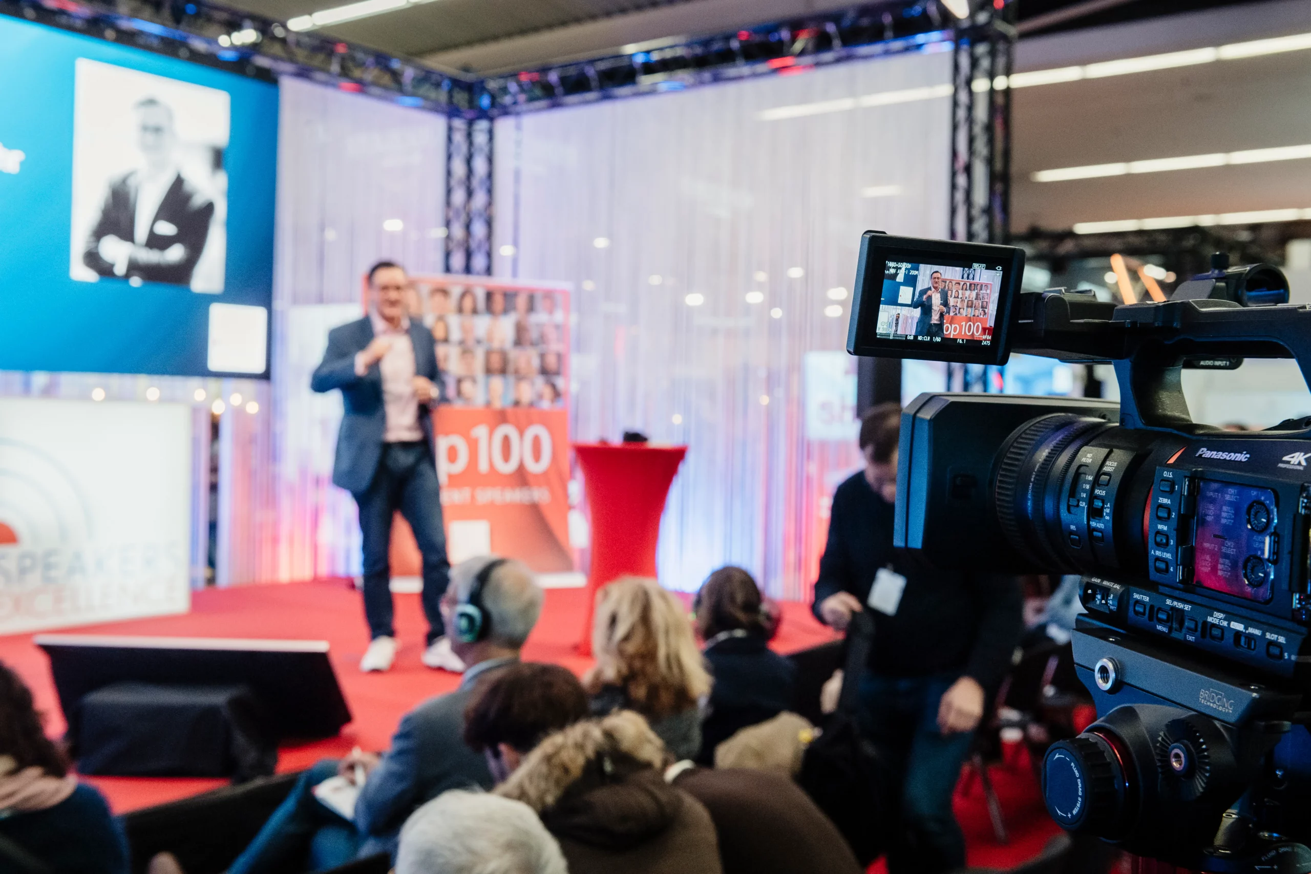 Eine Kamera filmt den Redner Dr. Nikolai A. Behr auf der Bühne einer Konferenz. Die Zuhörer sitzen auf Stühlen, einige mit Kopfhörern, während im verschwommenen Hintergrund ein Bildschirm mit dem Foto einer Person und einem Banner zu sehen ist. Die Szene ist lebendig und professionell.