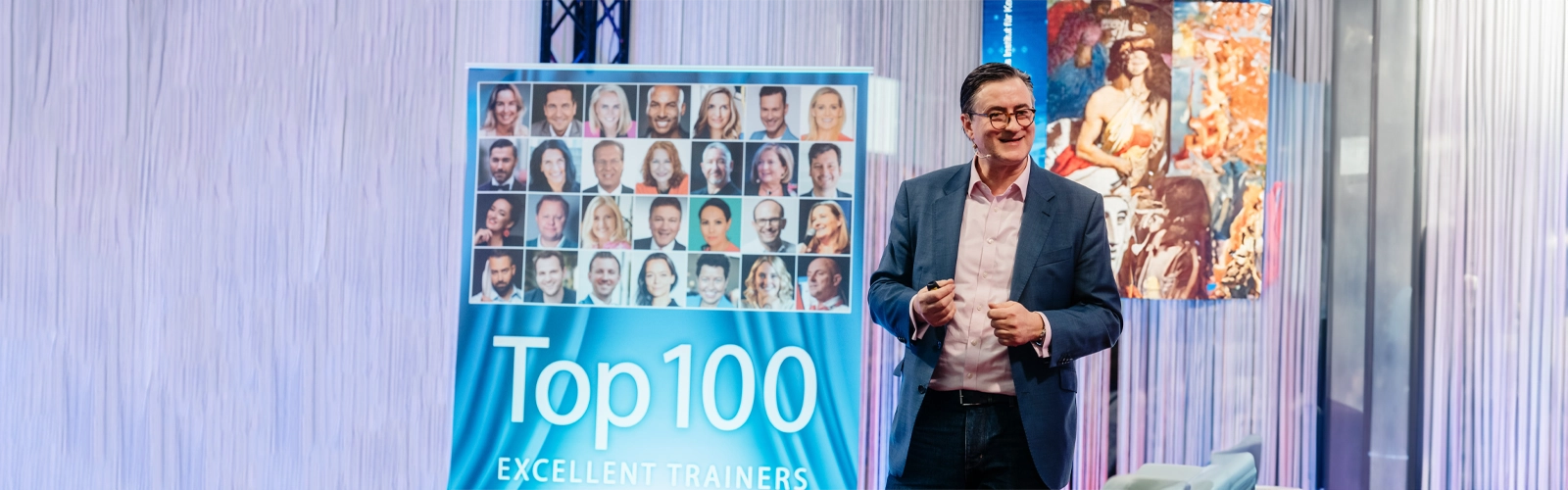 Medientrainer Dr. Nikolai A. Behr im Anzug steht auf der Bühne neben einem bunten Display, das Kopfbilder verschiedener Personen und den Text "Top 100 Excellent Trainers" zeigt. Im Hintergrund sind Vorhänge und ein Bildausschnitt eines Gemäldes zu sehen.