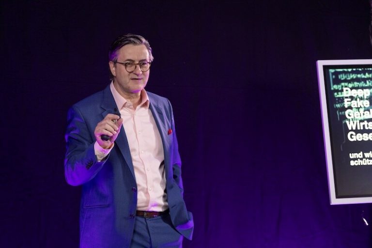 Ein Mann in einem blauen Anzug und mit Brille steht vor einem dunklen Hintergrund und gestikuliert, als würde er sprechen. Zu seiner Rechten befindet sich ein Bildschirm mit teilweise sichtbarem Text, der Fachbegriffe anzeigt. Violettes Licht beleuchtet die Szene.